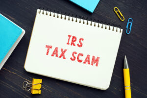 Notebook on a dark wooden surface with 'IRS TAX SCAM' stamped in red, surrounded by a blue wallet, colorful paper clips, and a yellow pen, symbolizing awareness about tax season scams for this Huff Insurance Blog
