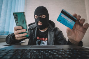 Man in a balaclava holding a credit card and smartphone, illustrating credit card fraud tactics, for Huff Insurance's blog on credit card fraud prevention